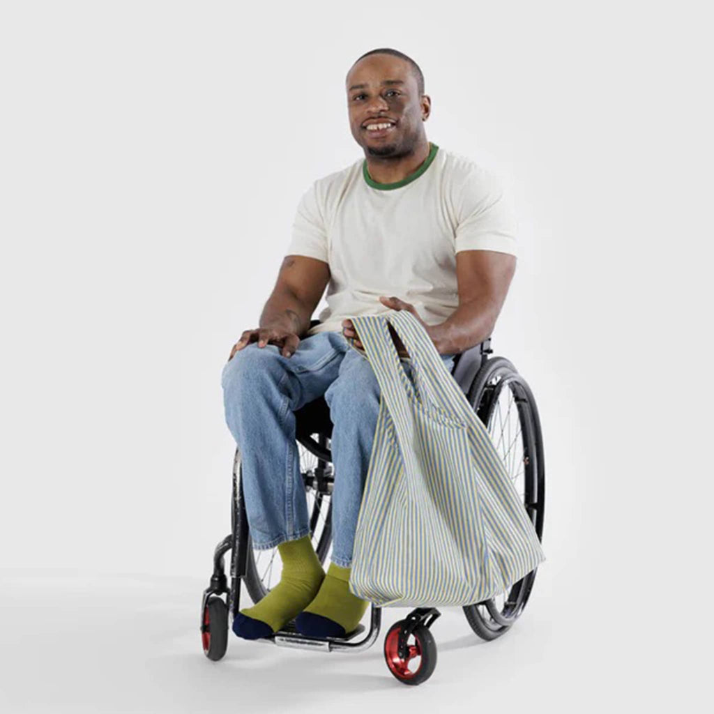 Baggu standard size eco-friendly recycled ripstop nylon reusable tote bag with yellow and blue thin stripe print, being held by a person in a wheelchair.