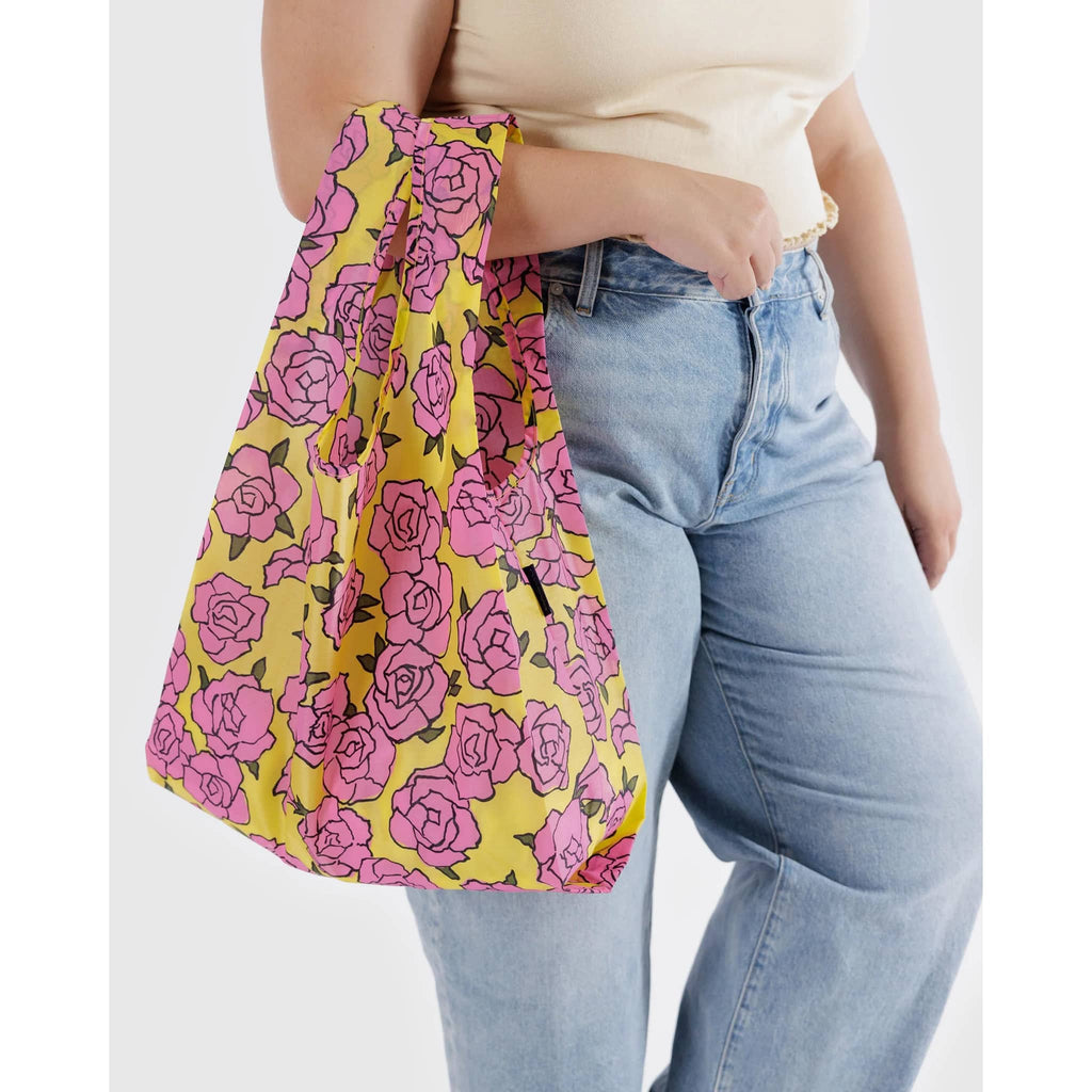 Baggu standard size eco-friendly recycled ripstop nylon reusable tote bag with a pink rose print on a yellow backdrop, on models arm.