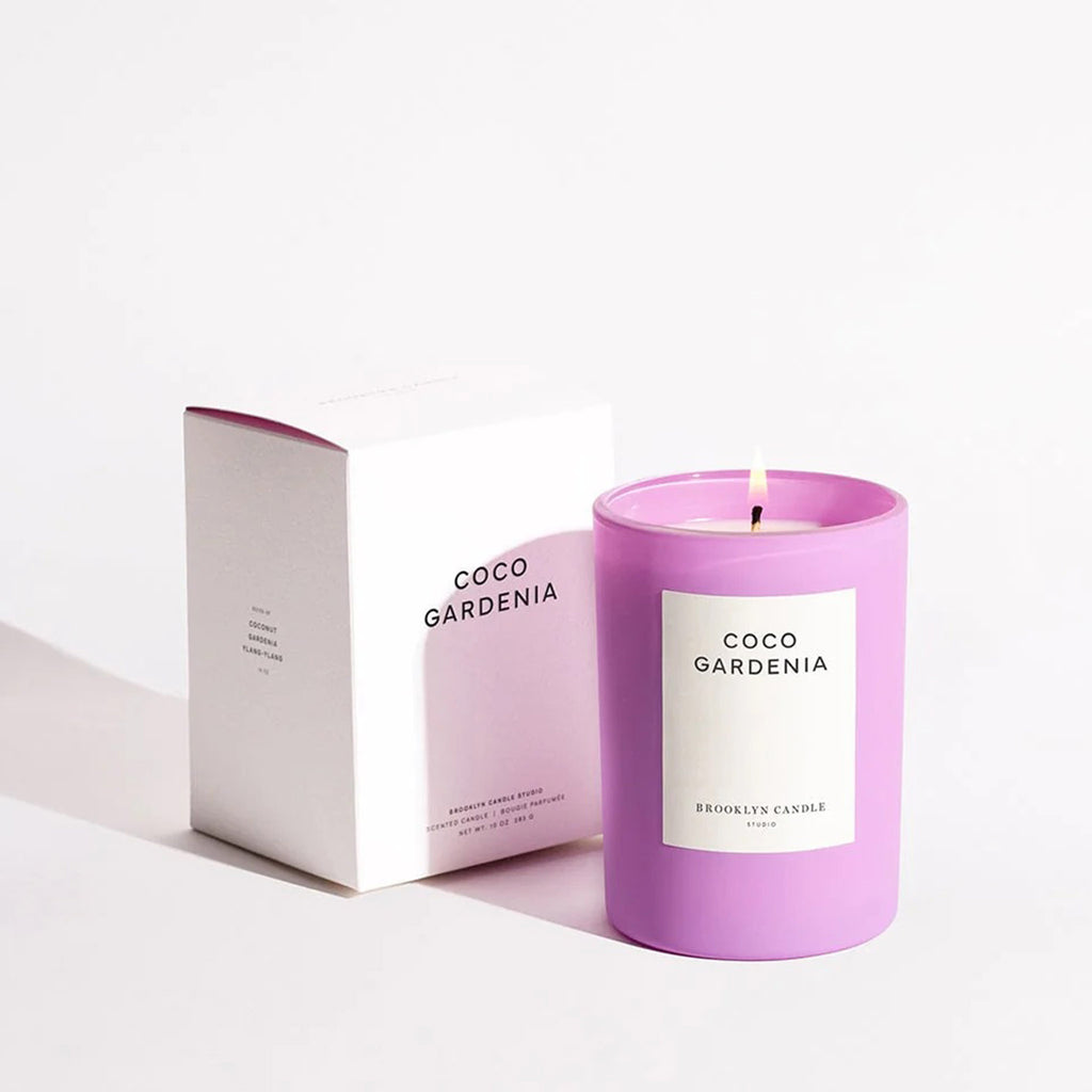Brooklyn Candle Studio Coco Gardenia scented candle in matte lilac glass vessel with white label and lit wick, beside white gift box.