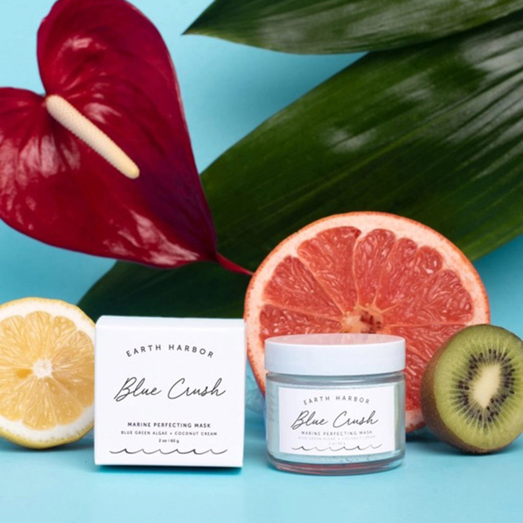 Earth Harbor Blue Crush Marine Perfecting Mask, blue powder mask in glass jar with white box packaging, a lemon, a grapefruit, a kiwi and a tropical flower on a blue background.