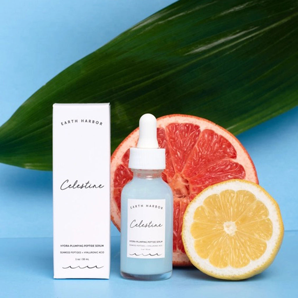 Earth Harbor Celestine Hydra-Plumping Peptide Serum in glass bottle with eyedropper, front view, with white box packaging and a grapefruit, lemon and tropical leaf on a blue background.