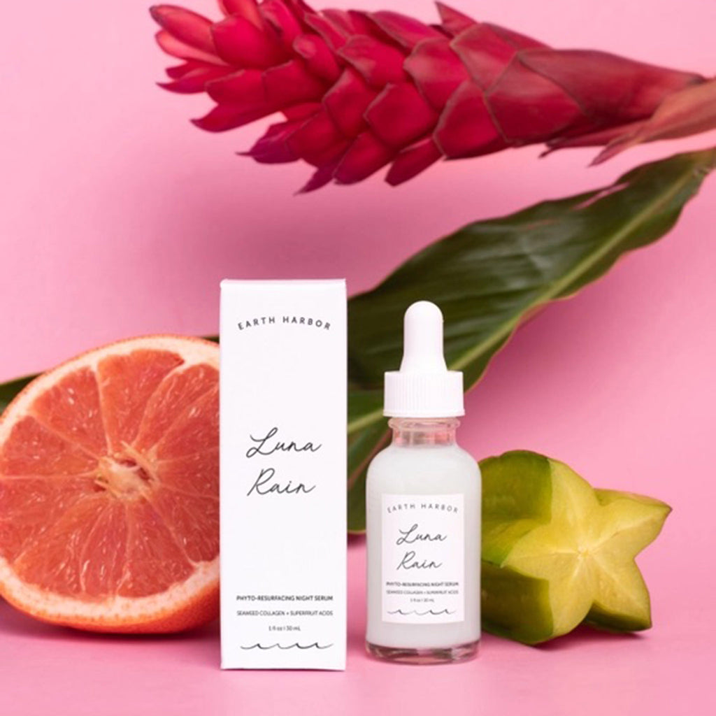 Earth Harbor Luna Rain Phyto-Resurfacing Night Serum in glass bottle with eyedropper, front view, with white box packaging and a grapefruit, star fruit and a tropical flower on a pink background.
