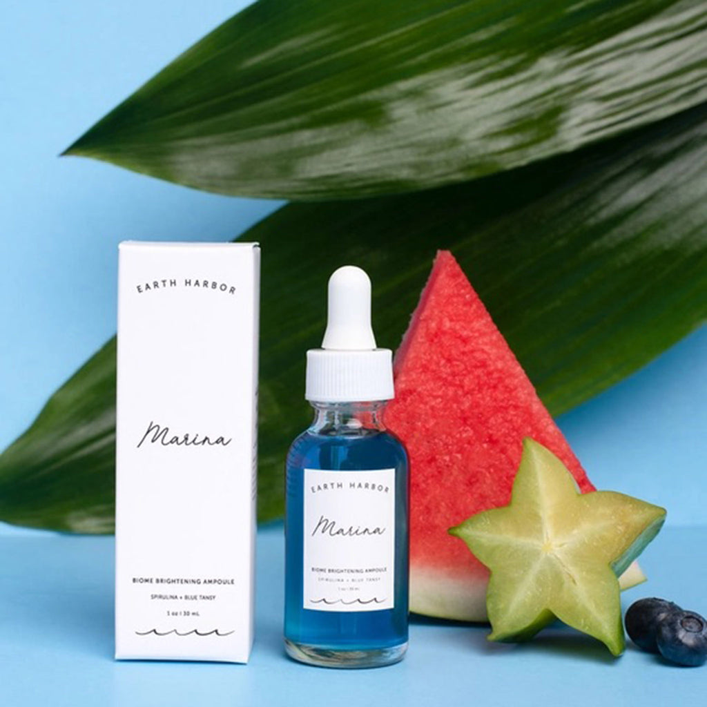Earth Harbor Marina Biome Brightening Ampoule Elixir in clear glass bottle with white box packaging, blueberries, starfruit, watermelon wedge and tropical leaves on a blue background.