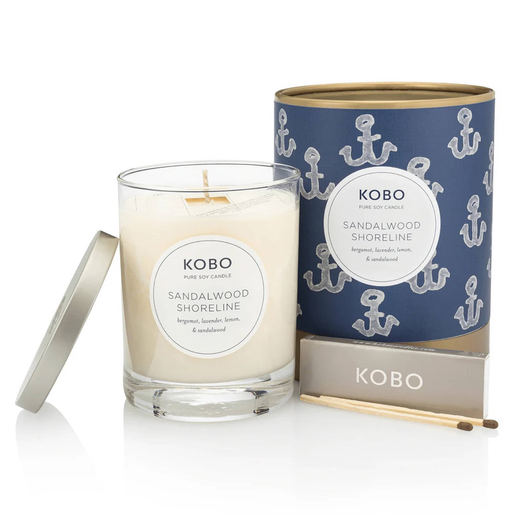 KOBO 11 ounce Sandalwood Shoreline candle, part of the Coastal Collection, in clear glass vessel with metal lid next to blue and white anchor illustrated canister packaging with a pack of wooden matches.