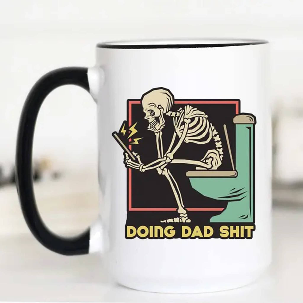 Mugsby white ceramic mug with black handle and rim with an illustration of a skeleton sitting on a toilet reading a tablet and "doing dad shit" in yellow lettering.