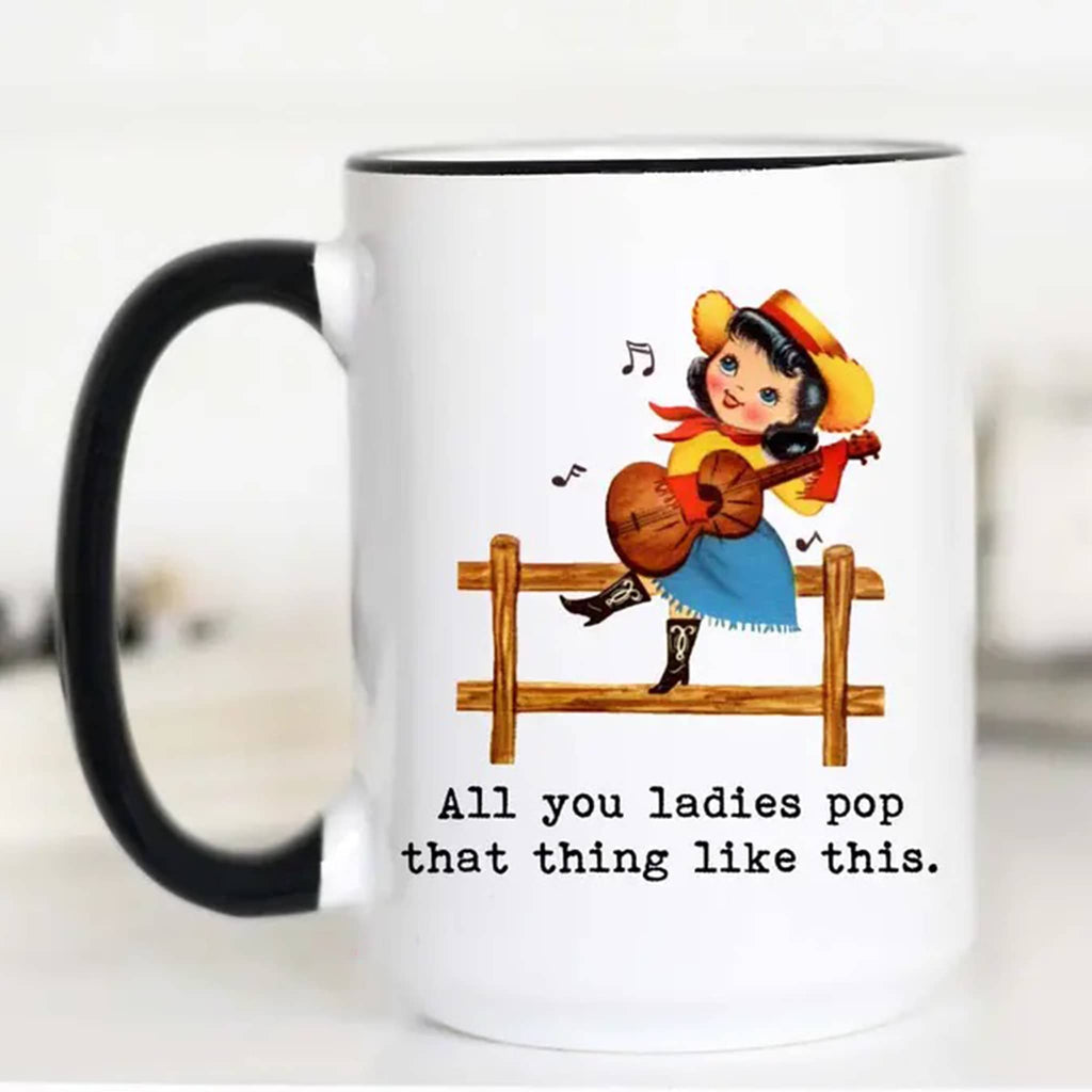 Mugsby white ceramic mug with black handle and rim with a vintage-inspired illustration of a girl in western wear playing a guitar and sitting on a wood fence with "all you ladies pop that thing like this" in black lettering.