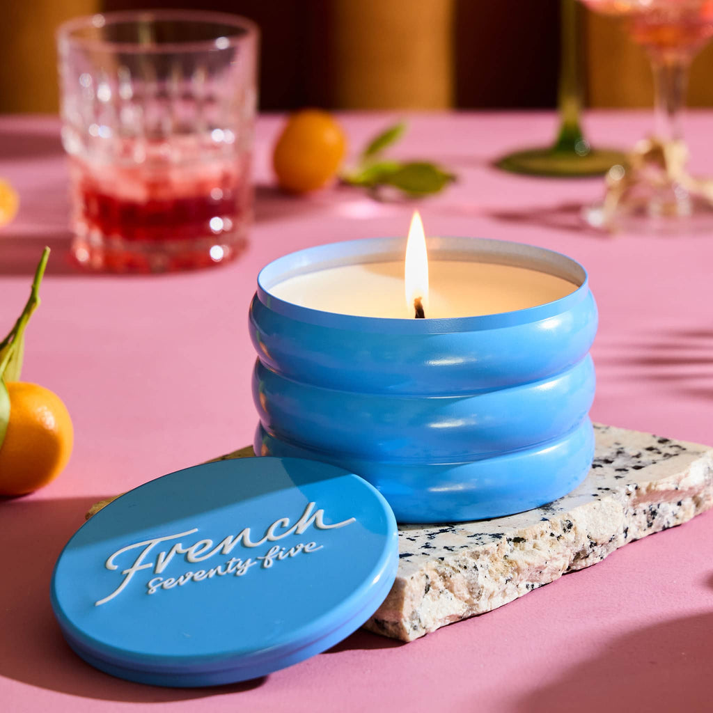 Rewined 13 ounce French 75 cocktail scented candle in blue ridged tin, lid off and wick lit with fruit and cocktail glasses in the background on a pink surface.