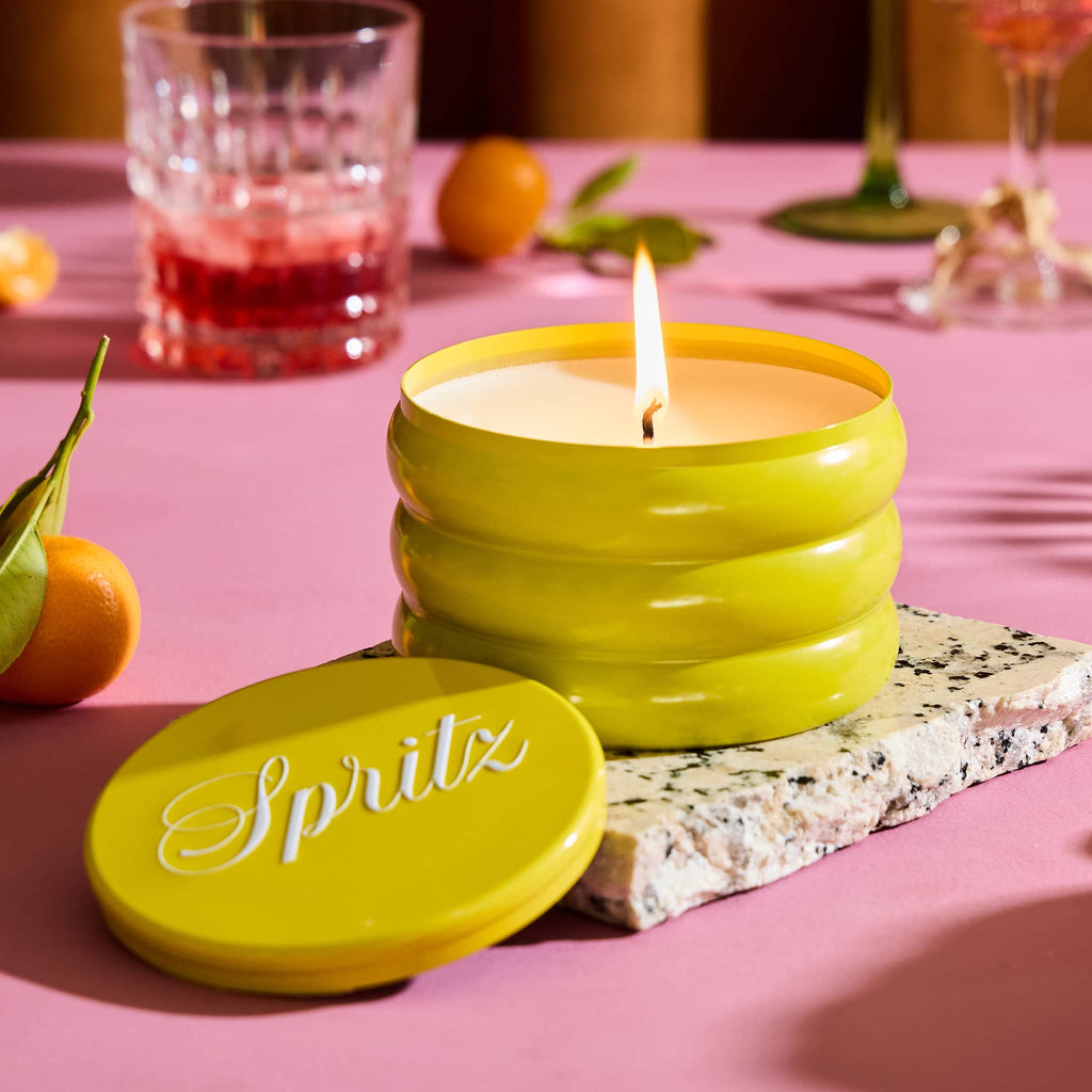 Rewined 13 ounce Spritz cocktail scented candle in yellow ridged tin, lid off and wick lit with fruit and cocktail glasses in the background on a pink surface.