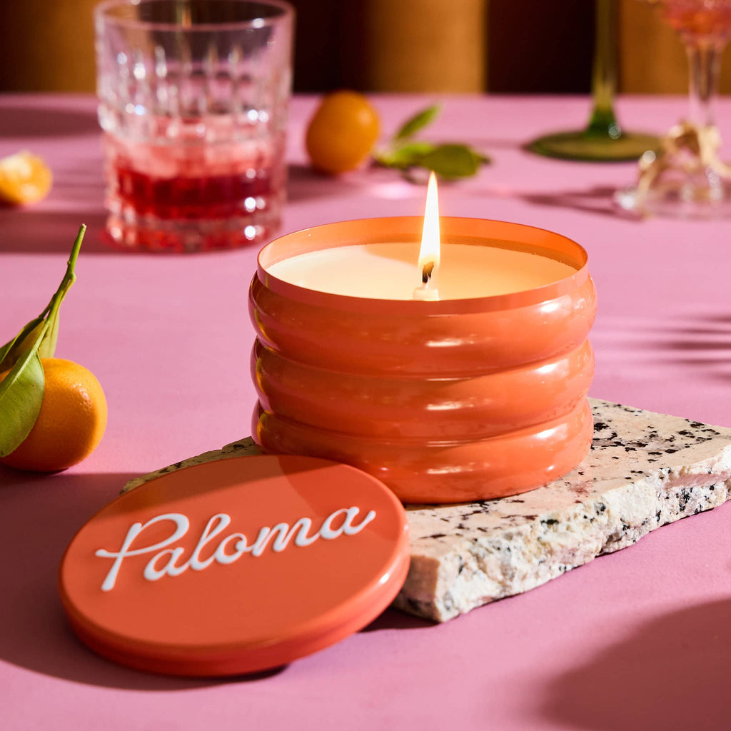 Rewined 13 ounce Paloma cocktail scented candle in orange ridged tin, lid off and wick lit with fruit and cocktail glasses in the background on a pink surface.