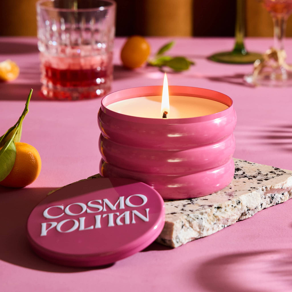 Rewined 13 ounce Cosmopolitan cocktail scented candle in pink ridged tin, lid off and wick lit with fruit and cocktail glasses in the background on a pink surface.