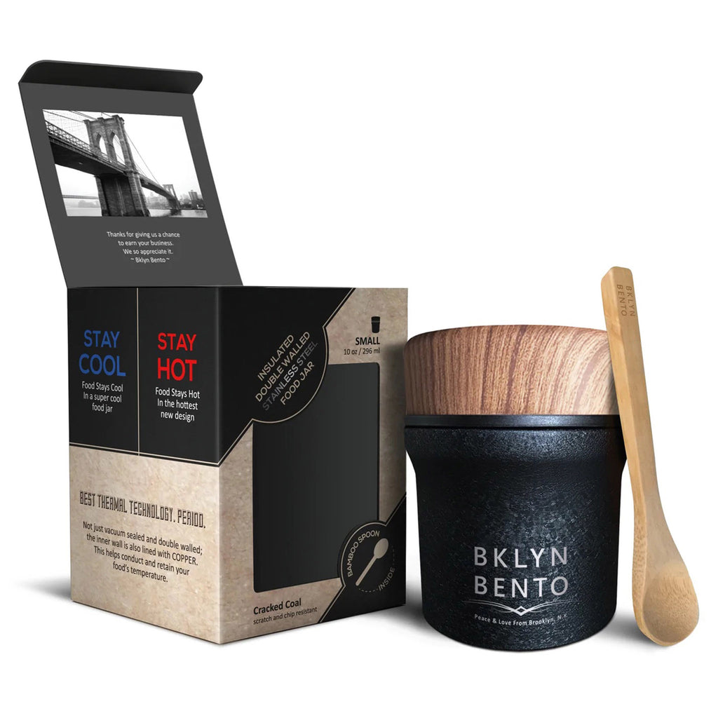 Black coated stainless steel insulated food jar with faux wood lid, a bamboo spoon is propped up against the jar and it's beside the tan and black paper box it comes in.