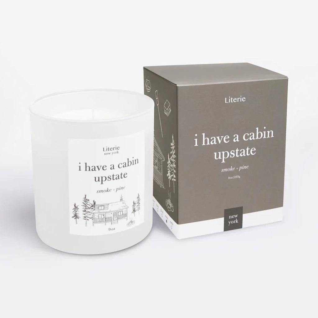 "I Have a Cabin Upstate" smoke and pine scented candle by Literie in a white matte glass tumbler with name of candle and cabin in the woods illustration in green and matching gray gift box with cabin in the woods illustration in white on the side.