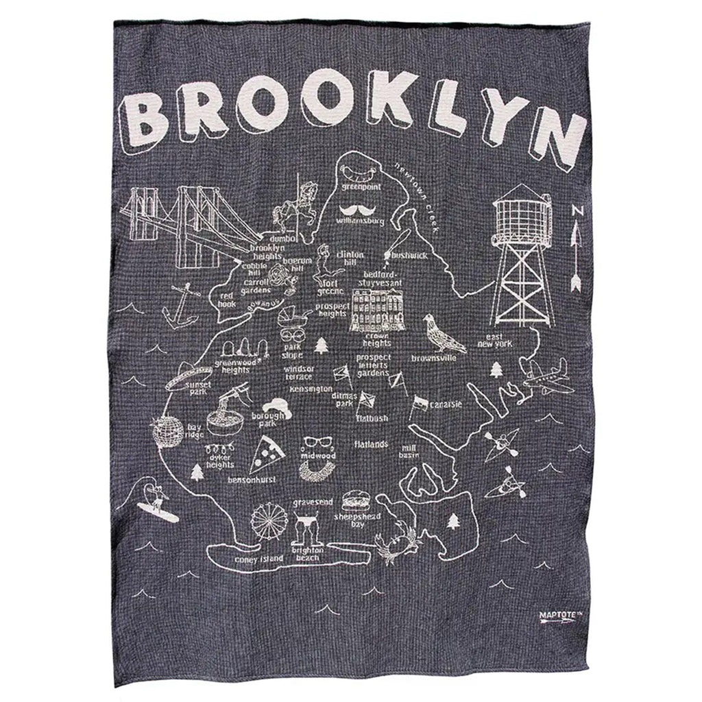 Overall of knit throw blanket with "brooklyn" at the top, the outline of the borough with neighborhood names and illustrations of various Brooklyn icons in white on a navy blue backdrop.