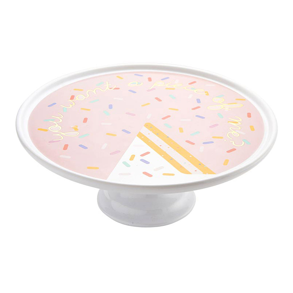 White ceramic cake stand with "you want a piece of me?" in yellow script lettering on the top on a pastel pink backdrop with colorful pastel sprinkles, side view showing top and pedestal.