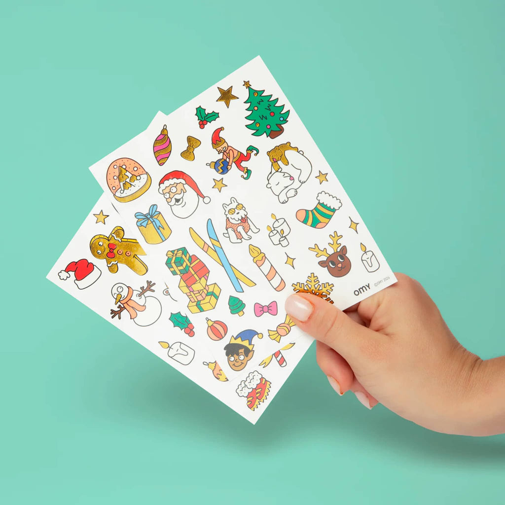Hand holding 2 sheets of Christmas-themed temporary tattoos against a green background.