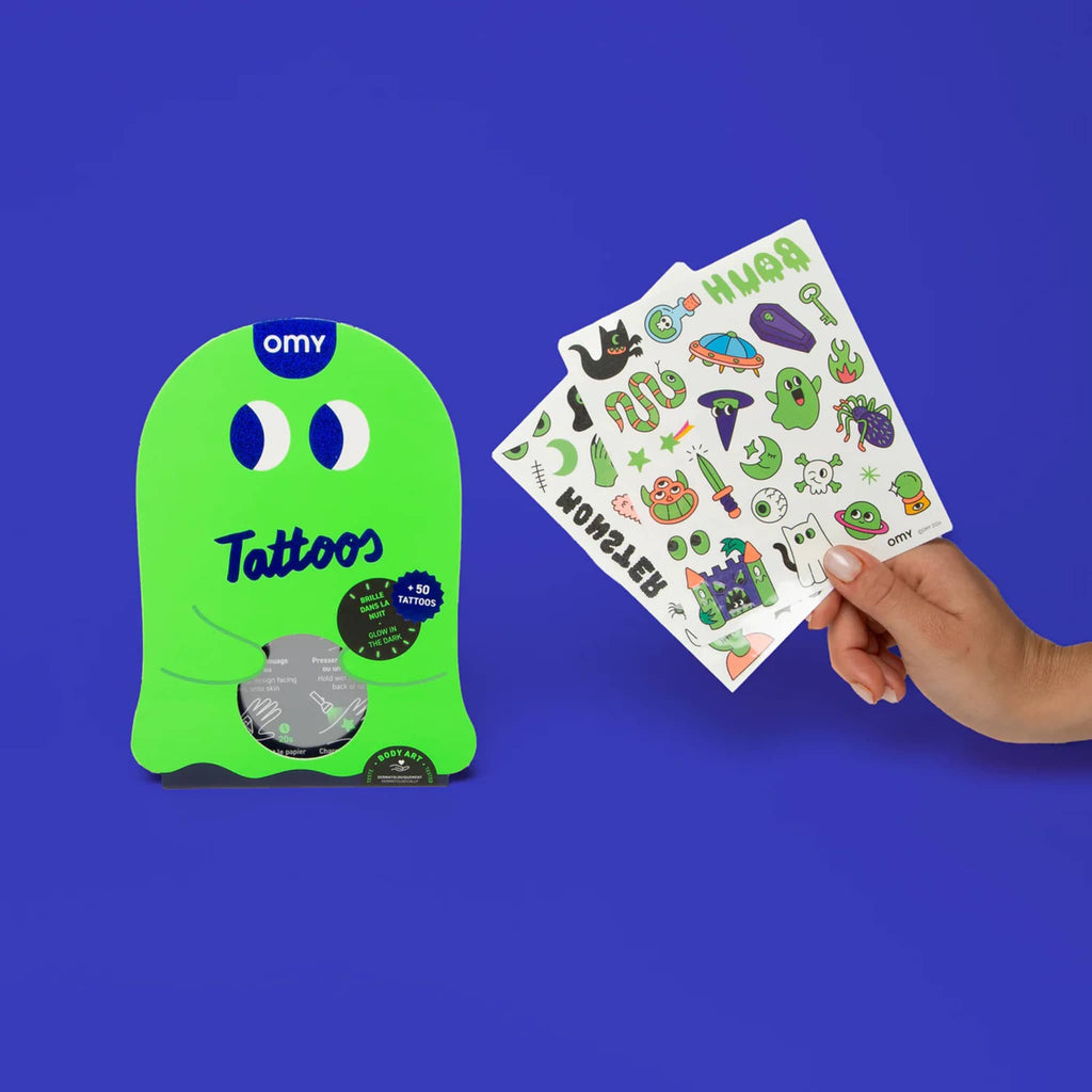 Green ghost shape packaging of OMY Halloween glow in the dark temporary tattoos beside a hand holding 2 sheets of tattoos beside it, on a blue background.