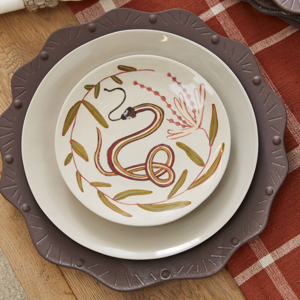 Accent Decor Parable Snake Ceramic Plate with a hand-painted red, orange and green snake and leaf design on a cream backdrop, plate is shown with a larger plate and charger underneath it in a place setting.