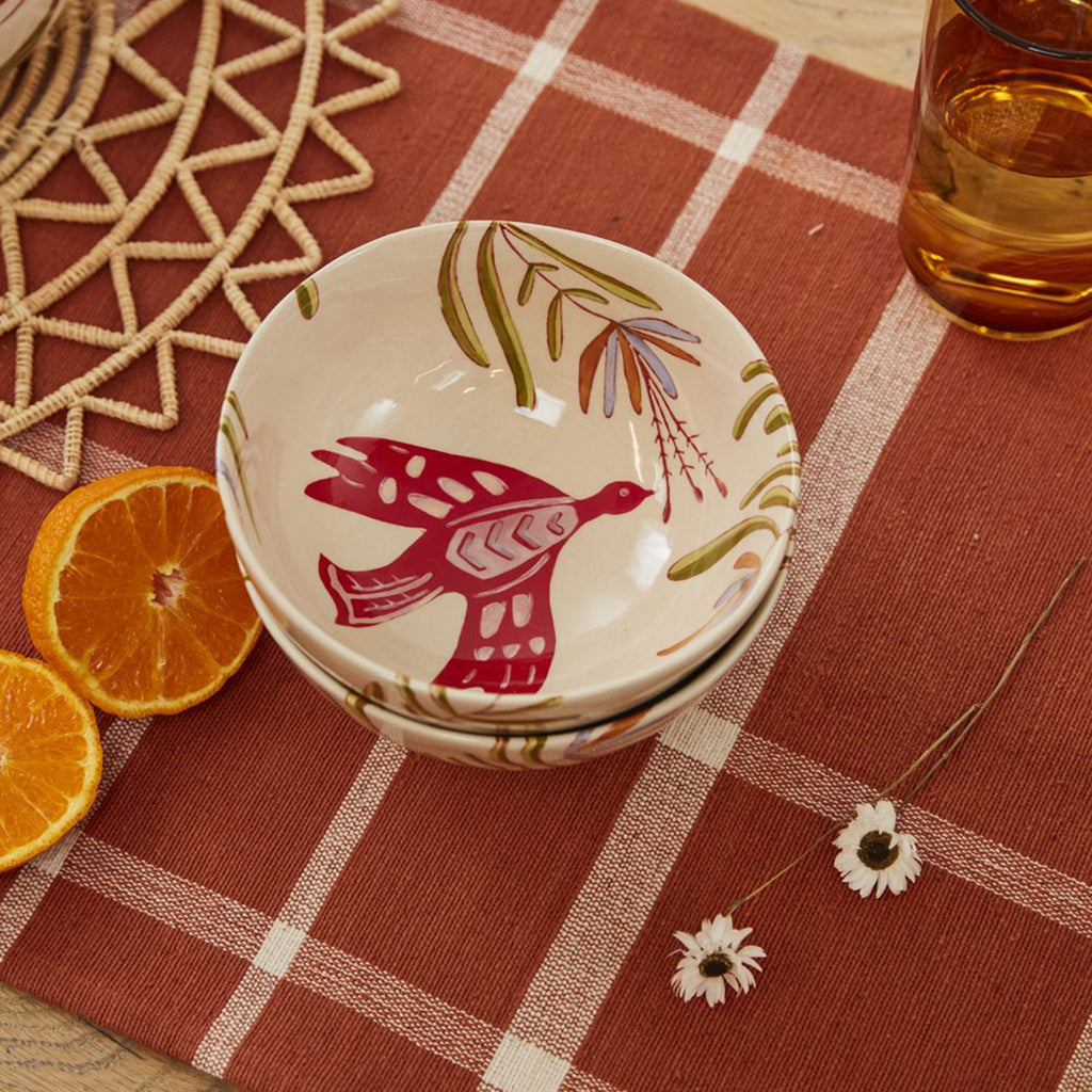 Accent Decor Parable Bird Ceramic Bowl with a hand-painted red, orange and green bird and leaf design on a cream backdrop, bowl is shown from overhead as a stack of 2 on a rust colored table runner.