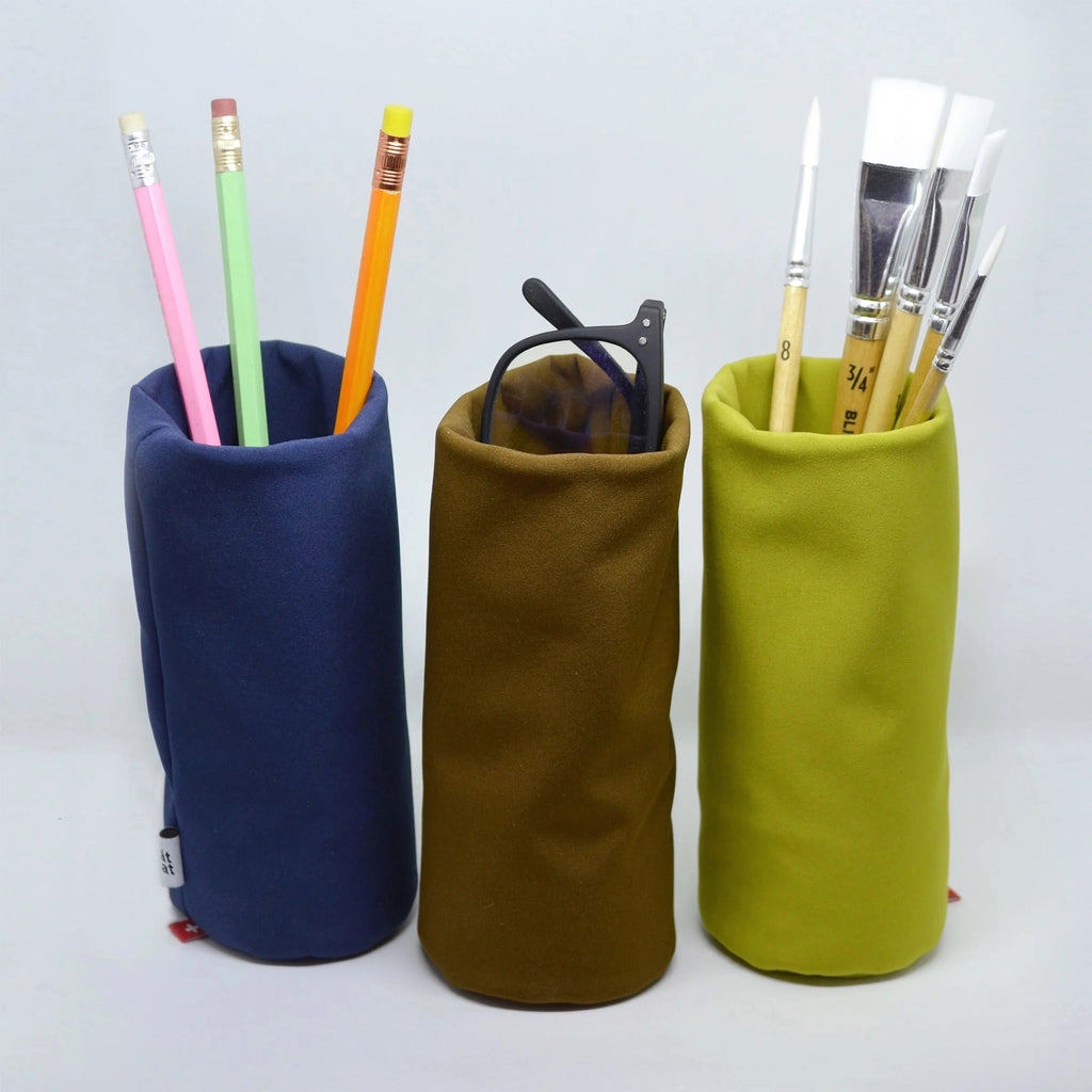 Ameico Tat Tat Sacco Multi-Purpose soft-sided stand-up storage pouches in dark blue (with pencils), brown (with eyeglasses) and chartreuse (with paintbrushes).
