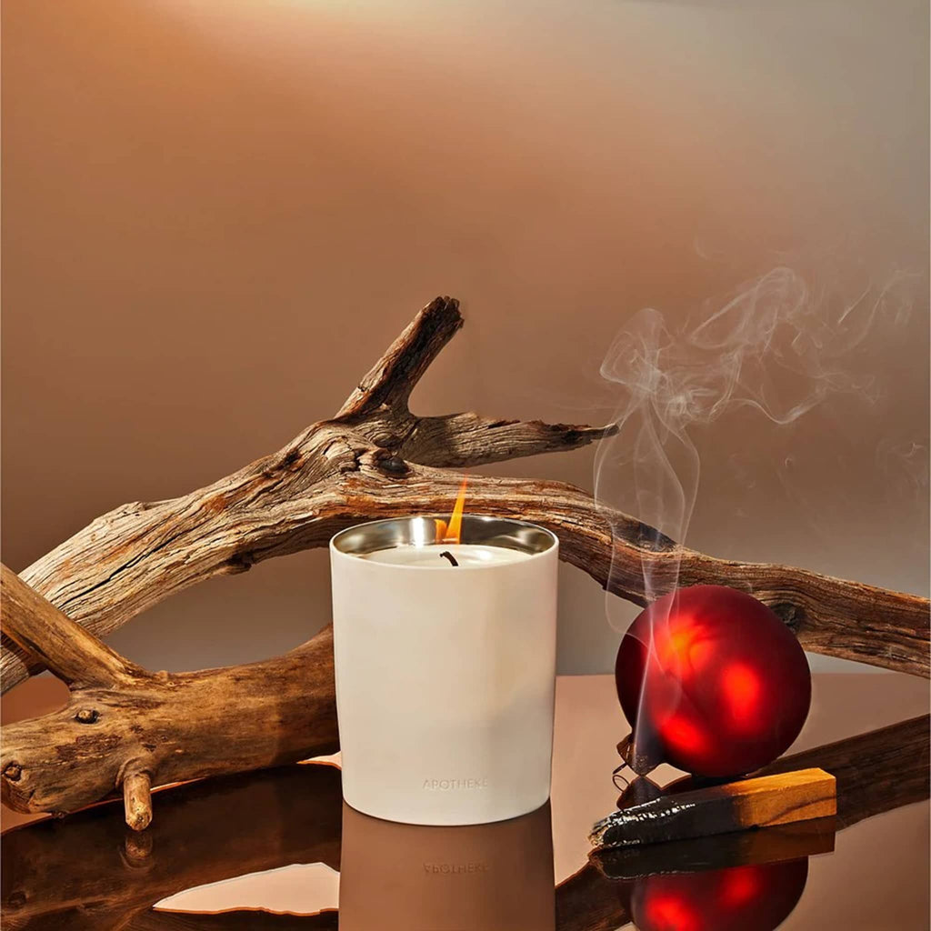 Apotheke Firewood 12.5 ounce scented candle in white ceramic vessel with gold interior, shown with tree branches, a smudge stick and a red ornament.