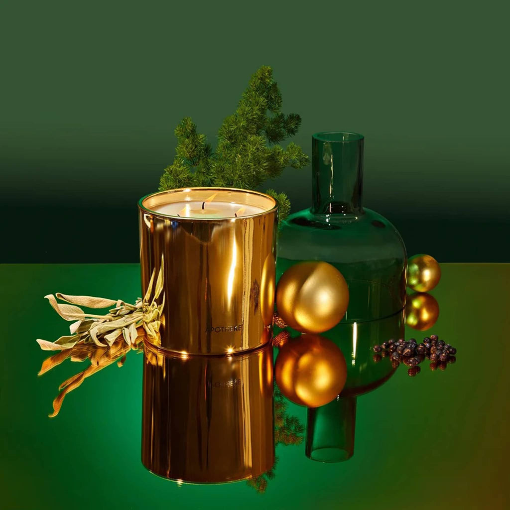 Apotheke Black Cypress 26 ounce 3-wick scented candle in gold vessel, shown on a green mirrored surface with botanicals, gold ornaments and a green glass decanter.