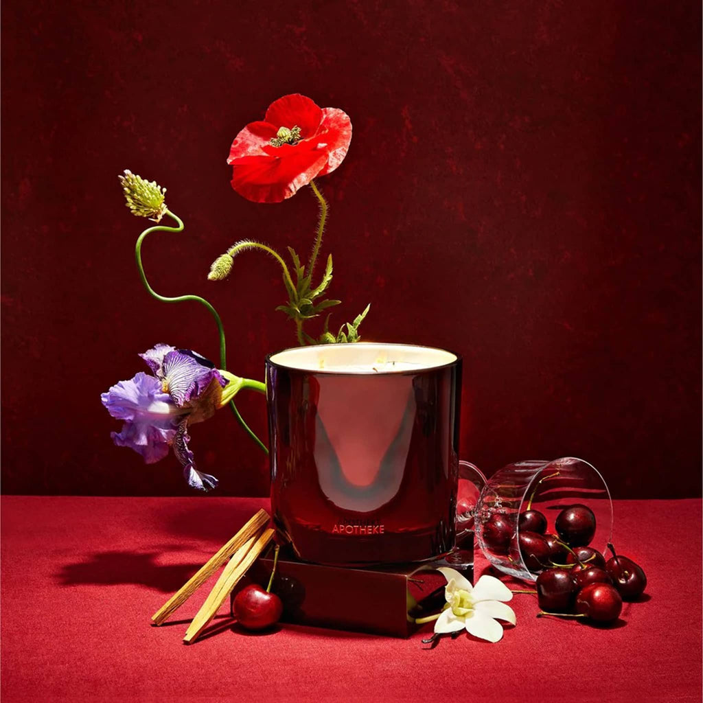 Apotheke 26 ounce Bitter Cherry scented soy wax blend candle in deep cherry red glass vessel with gold interior and 3 wicks, lit and surrounded by flowers and cherries with a deep red background.