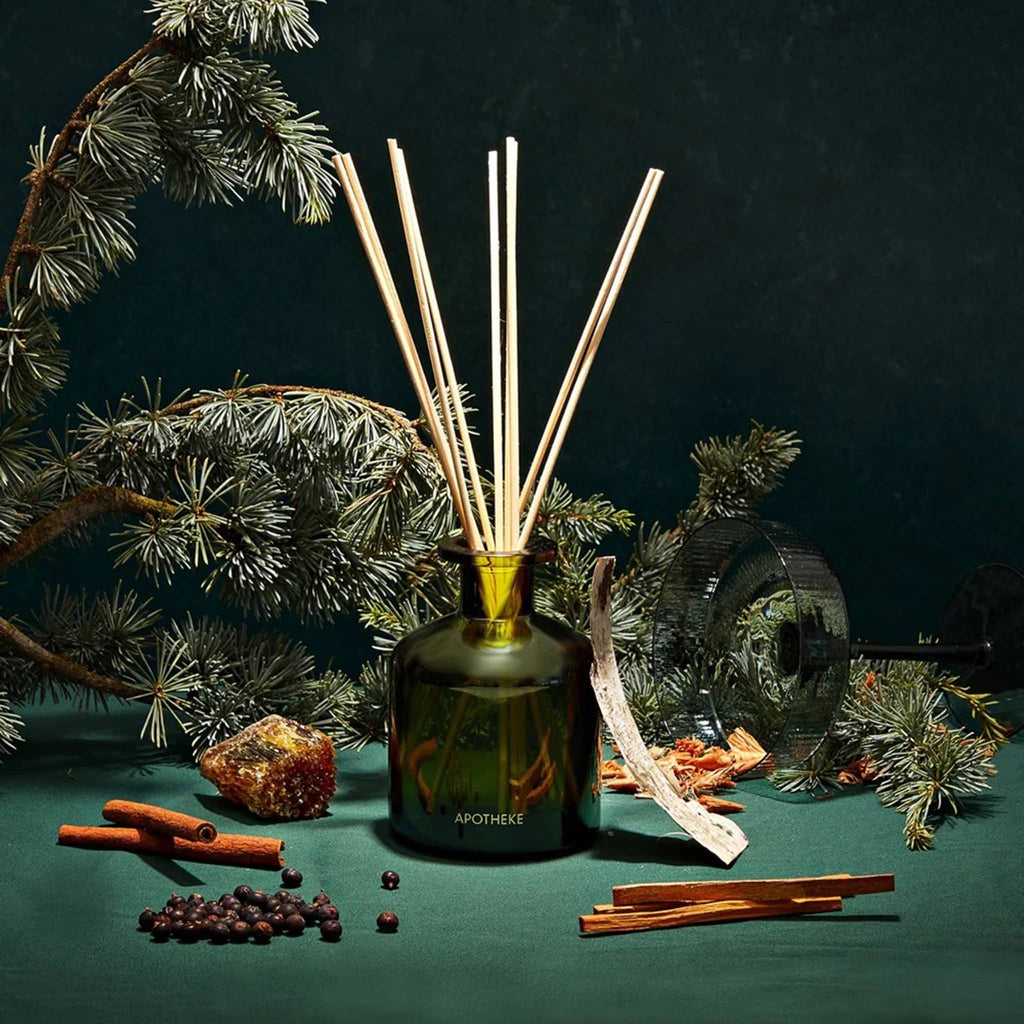 Apotheke Juniper Birch scented limited edition reed diffuser in dark green glass bottle with natural reeds on a green background with botanicals and a green coupe glass.