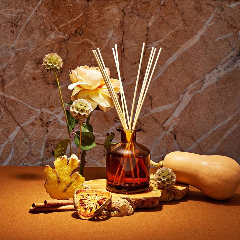 Apotheke 6.7 ounce Pumpkin Ginger scented reed diffuser with sticks in bronze glass bottle, surrounded by flowers and fruit props.