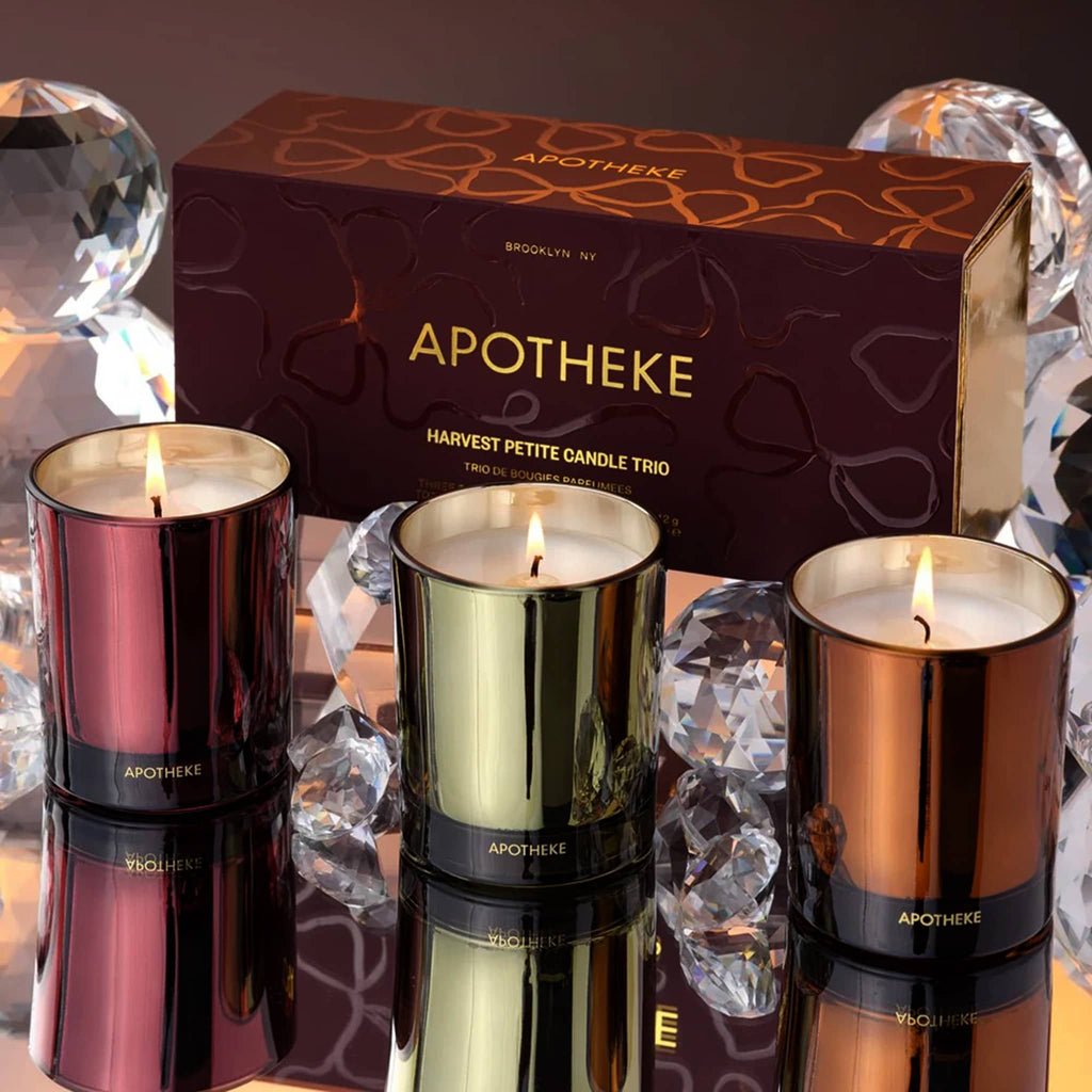 Three lit candles in metallic red, green, and orange glass vessels with an 'Apotheke' branded box in the background.