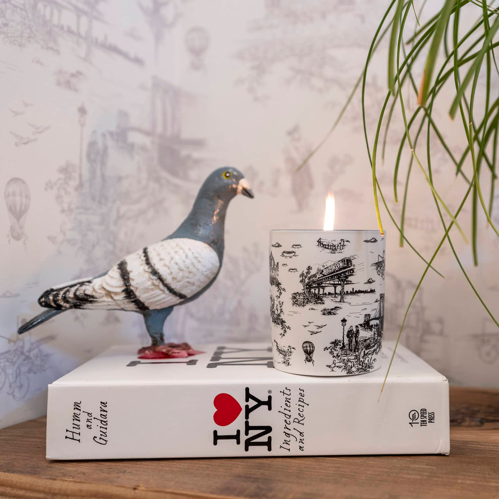 Apotheke x Flavor Paper Charcoal scented candle in white ceramic vessel with the Brooklyn Toile print in black, lit and sitting next to a pigeon figurine on top of an I love NY book with the wallpaper in the background.