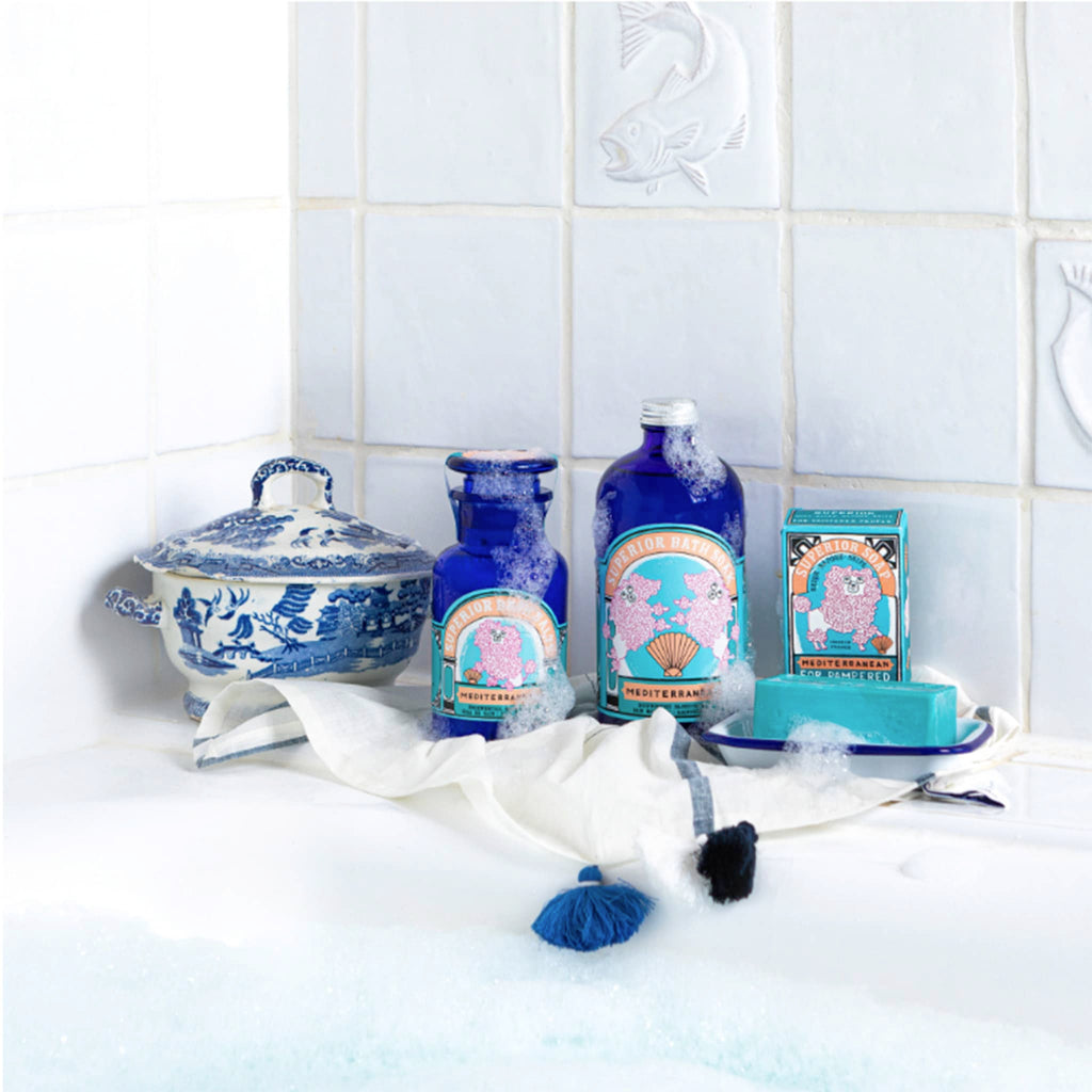 Archivist Gallery Mediterranean Bath Salts, Bath Soak and Hand Soap Bar in packaging on the ledge of a white bath tub filled with suds.