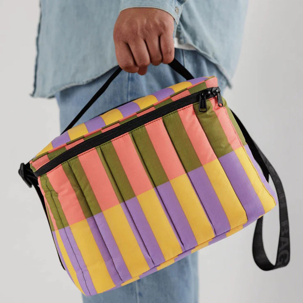 Baggu Insulated Puffy Cooler Bag with Sunset Quilt Stripe print in coral, olive green, yellow and purple with black zipper, shoulder strap and handle, being carried.