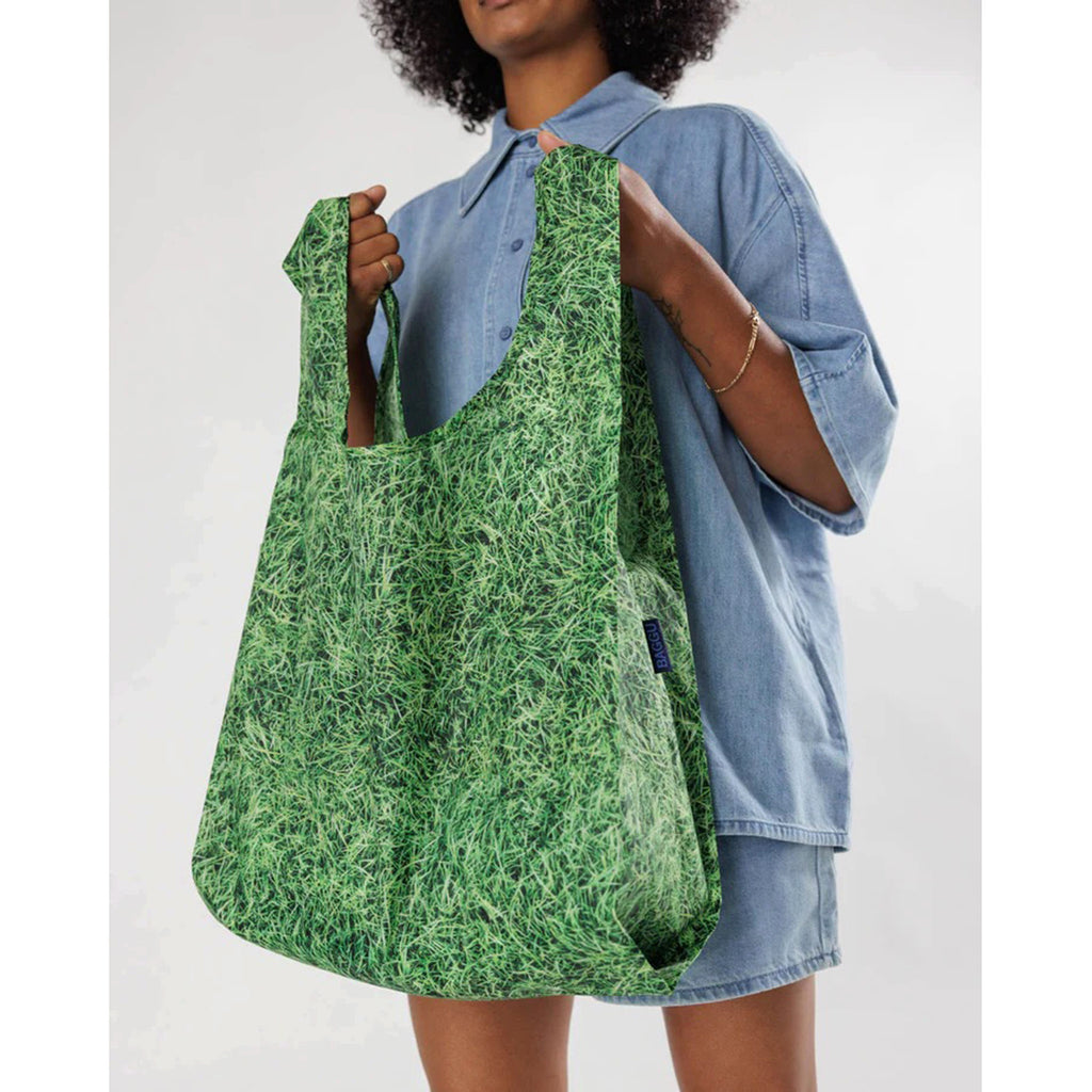 Baggu standard size eco-friendly-recycled ripstop nylon reusable tote bag with green grass pattern, held by handles in models hands.