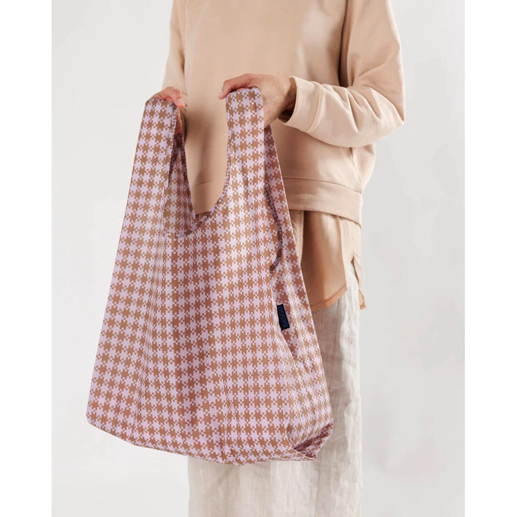 Baggu standard size eco-friendly-recycled ripstop nylon reusable tote bag with pink and brown pixel gingham pattern, being held by the handles.