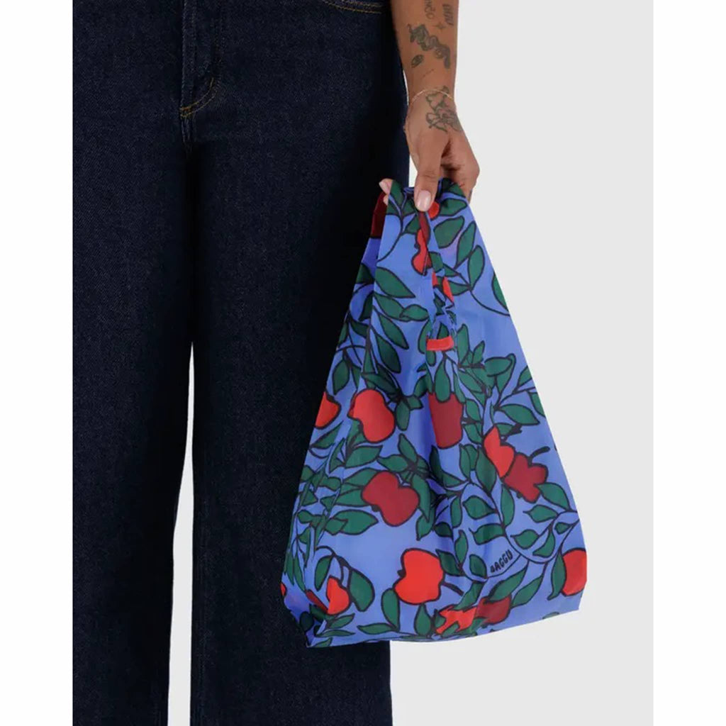 Blue bag with red apple and green leaf pattern held by a person wearing dark jeans.