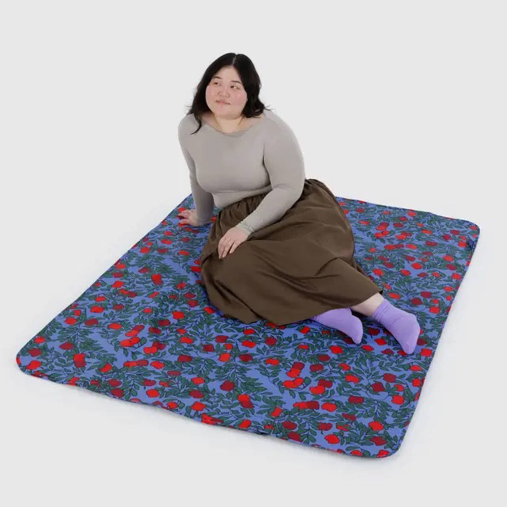 Woman sitting on a Baggu Apple Tree puffy recycled ripstop nylon and polyester picnic blanket.