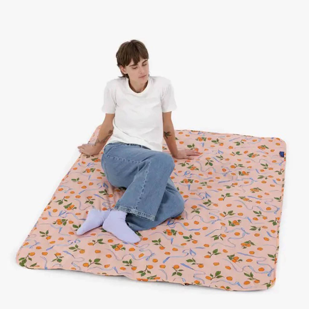 Baggu Puffy Picnic Blanket in Apricots and Ribbons print, flat with model lounging on it.