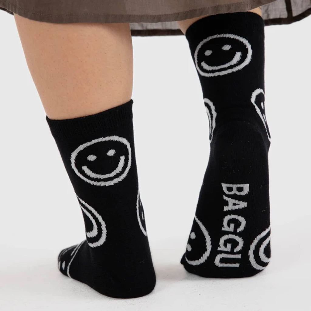 Baggu bamboo rayon unisex Happy crew socks with gray smiley faces on a black background, being worn by a model, back and sole view.