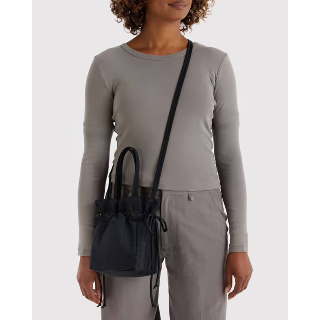 Woman with a black drawstring bag worn crossbody, wearing a gray long-sleeve top and light gray pants on a white background.