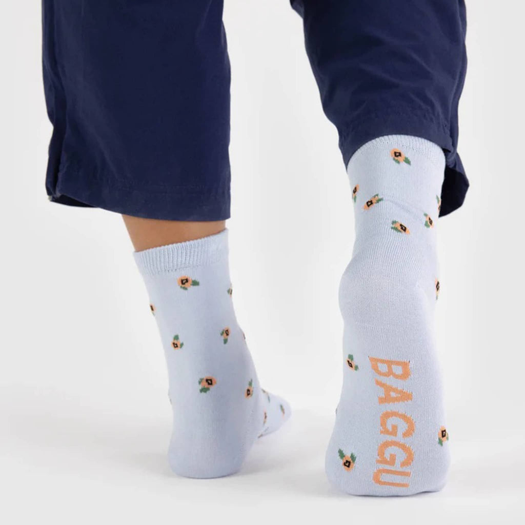 Baggu bamboo rayon unisex Rosette crew socks with small peach flowers on a pale blue background, being worn by a model, back and sole view.