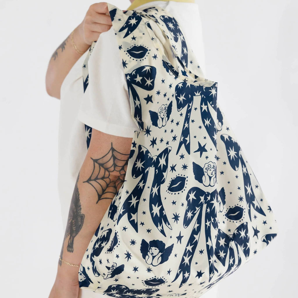 Baggu standard size eco-friendly recycled ripstop nylon reusable tote bag with blue and white Cherub Bows print, model holding it over their shoulder.