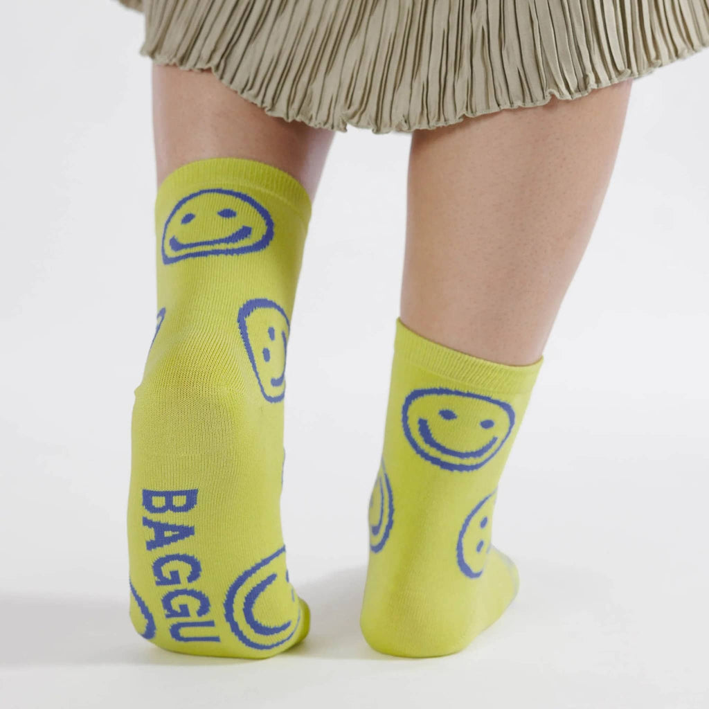 Baggu bamboo rayon unisex crew socks with blue smiley faces on a citron background, being worn by a model.