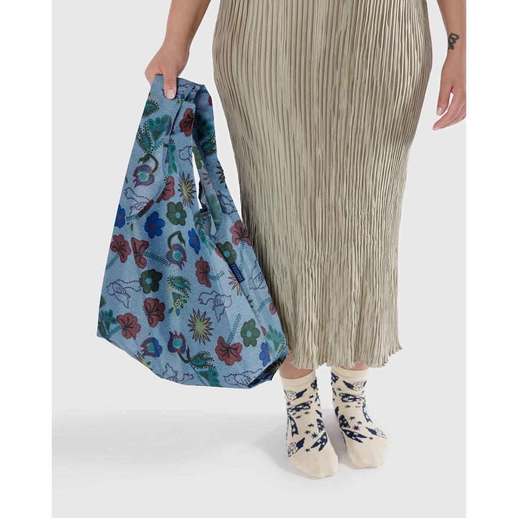 Baggu standard size eco-friendly recycled ripstop nylon reusable tote bag with floral and bird illustrations on denim colored backdrop, in model's hand.