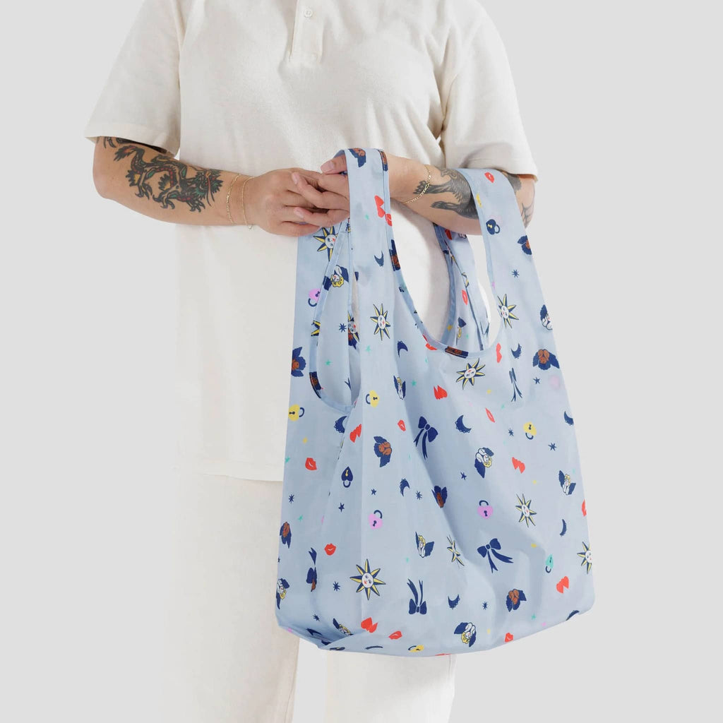 Baggu standard size eco-friendly recycled ripstop nylon reusable tote bag with Ditsy Charms print on a light blue backdrop, hanging on a model's arm.