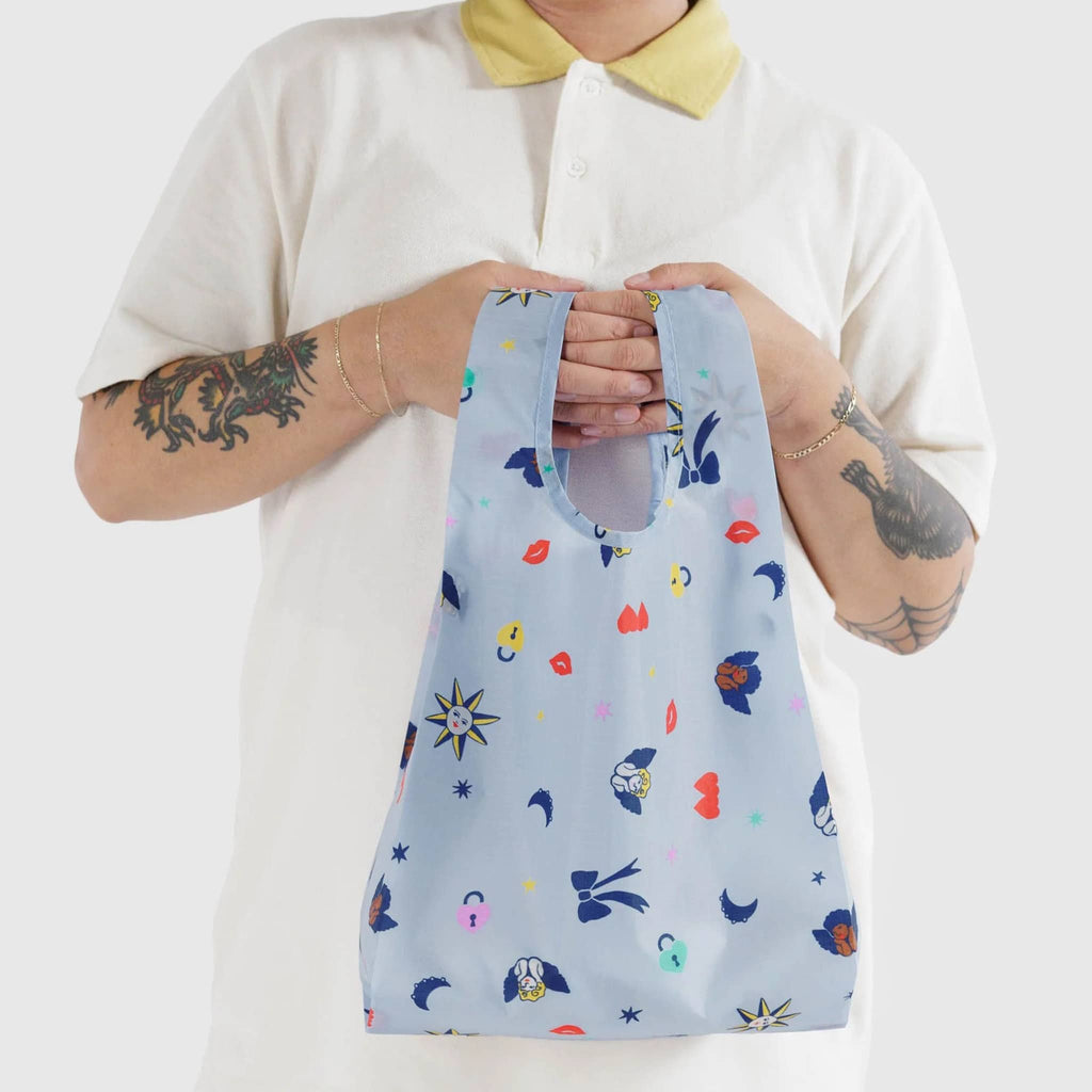 Baggu baby size eco-friendly recycled ripstop nylon reusable tote bag with Ditsy Charms print on a light blue background, model holding the bag in their hands.