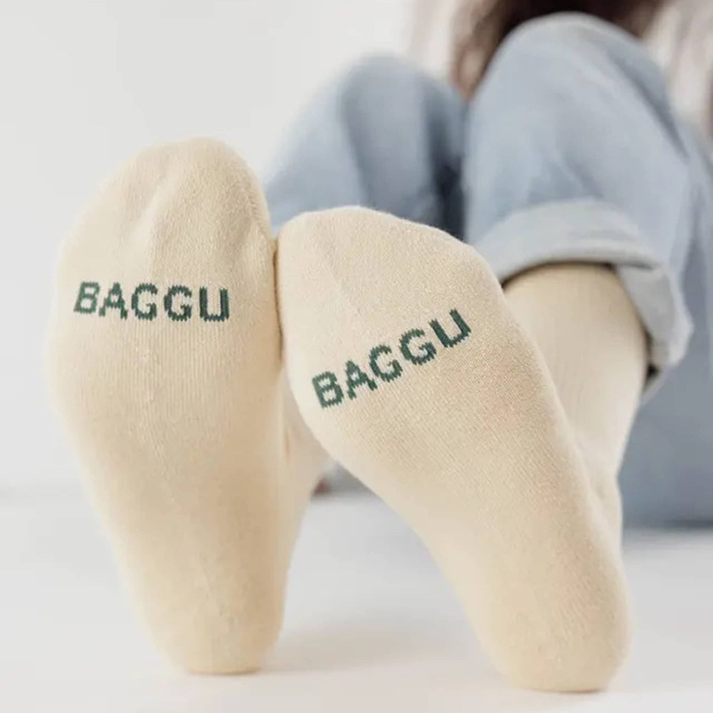Ribbed ecru socks with 'BAGGU' in dark green text worn by a person sitting on a white surface.