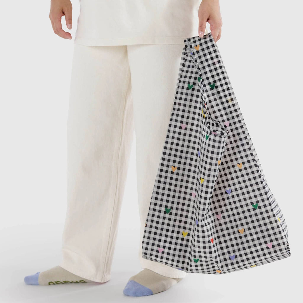 Baggu standard size eco-friendly recycled ripstop nylon reusable tote bag with tiny colorful hearts on a black and white gingham backdrop, in model's hand.
