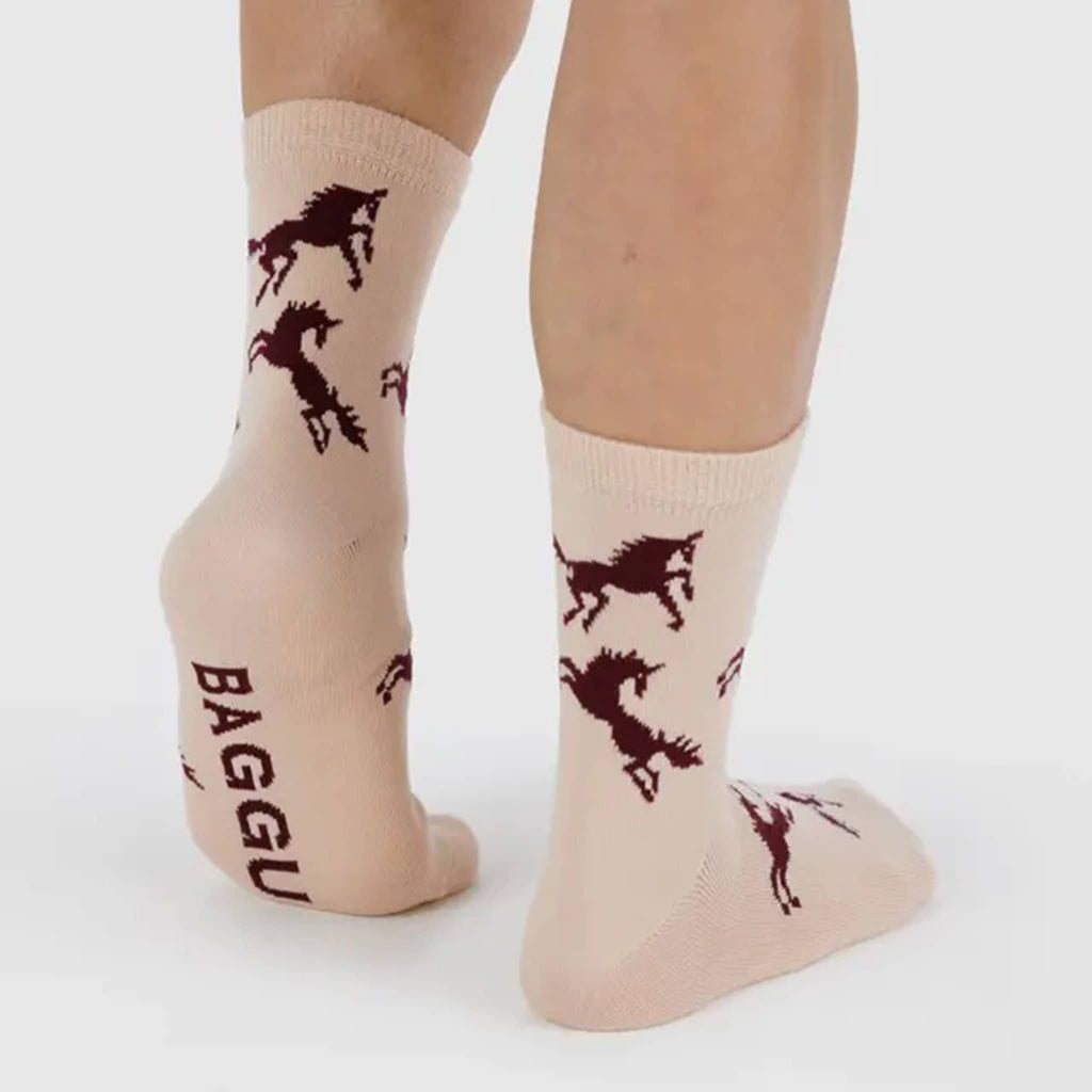 Pink socks with burgundy horse designs and 'BAGGU' branding on a persons feet, on a white background.