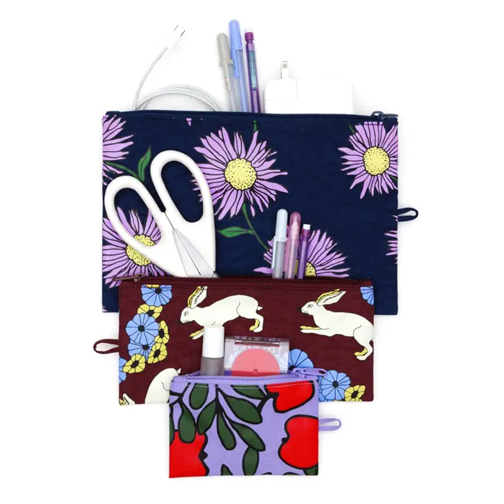 Baggu Night Orchard recycled heavyweight nylon flat zipper pouch, set of 3 in different prints, open with cosmetics, scissors, pens and a charger.