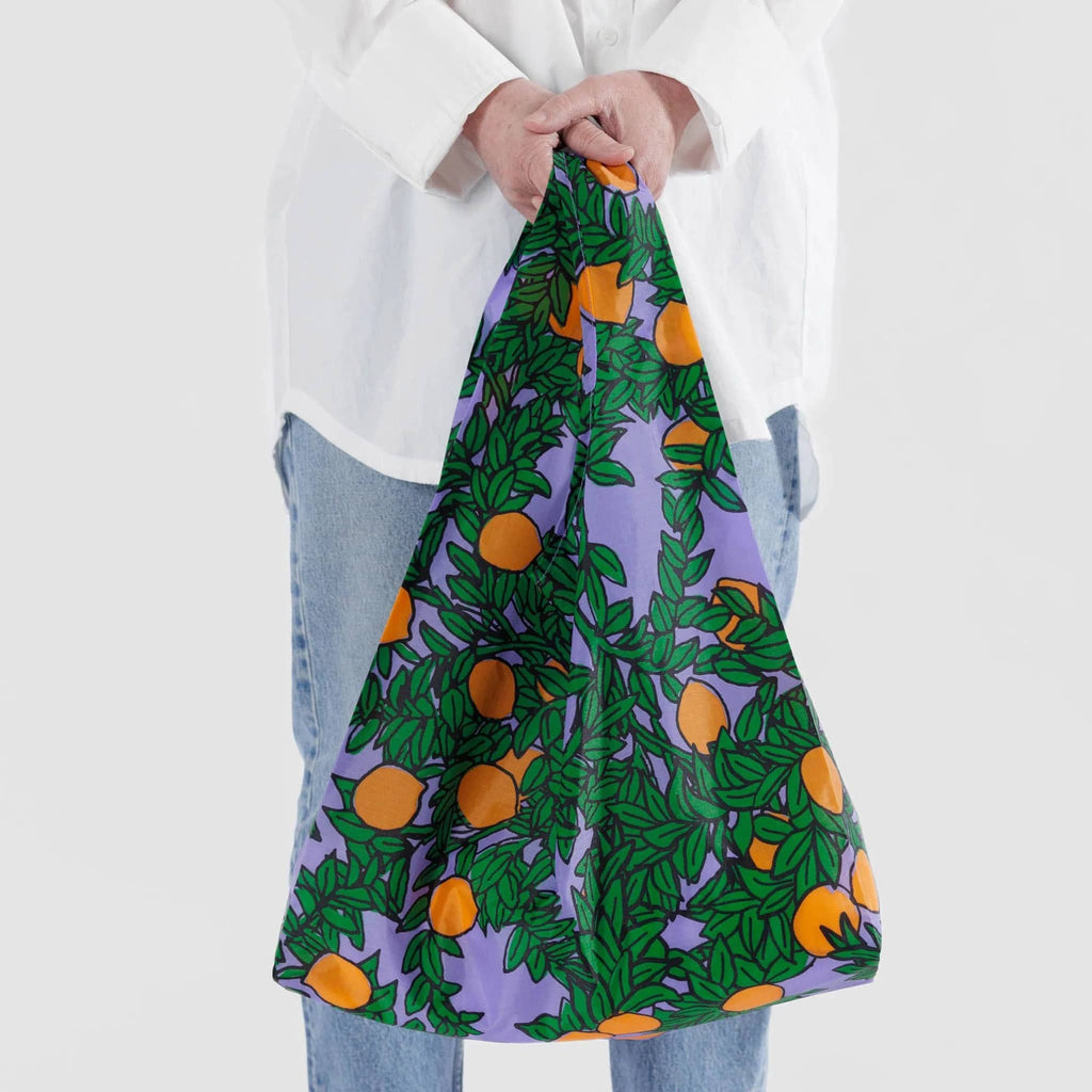 Baggu standard size eco-friendly recycled ripstop nylon reusable tote bag with an orange tree print on a periwinkle backdrop, in models hands.